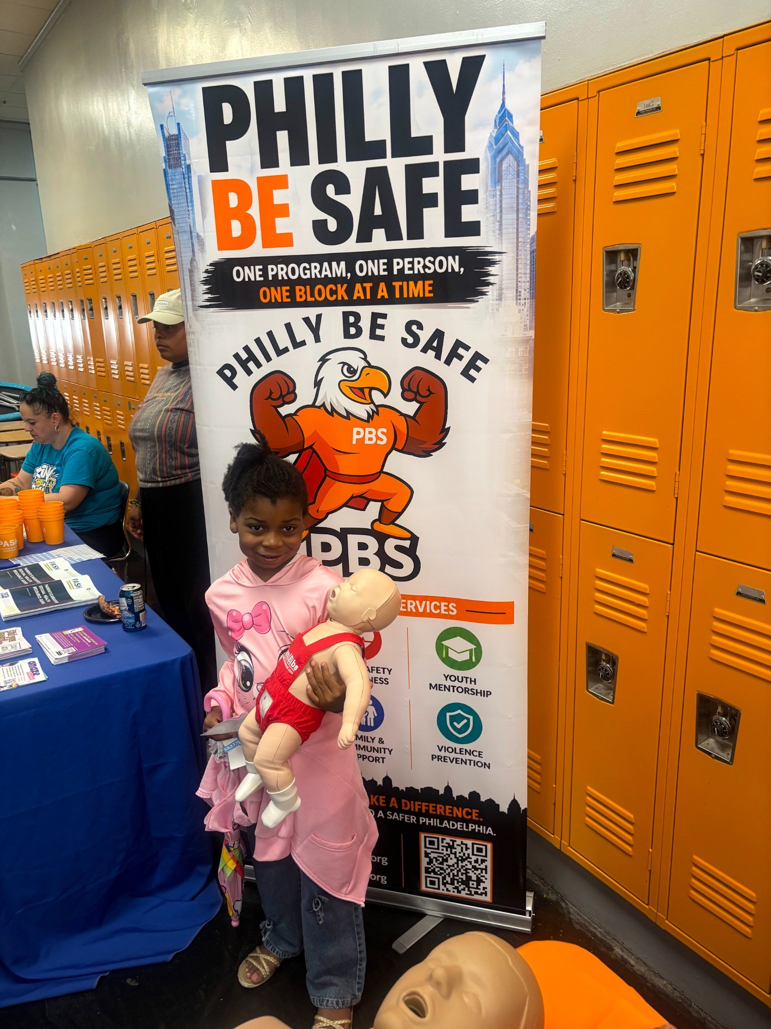 Child holding a CPR training manikin in front of the Philly Be Safe community preparedness banner